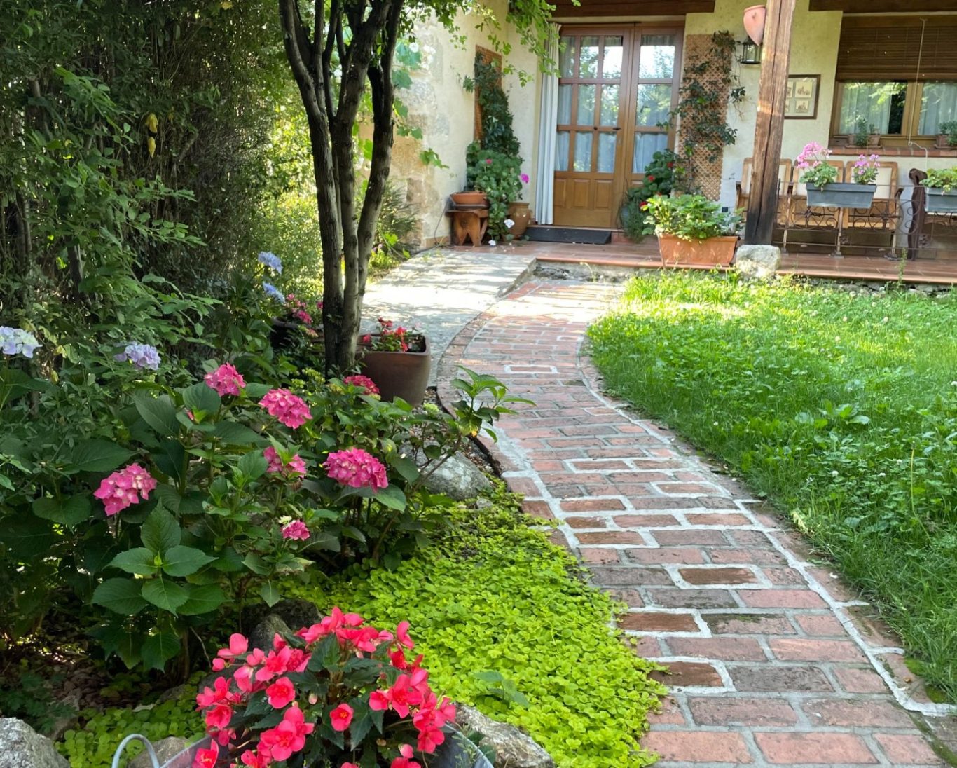 Porche con acceso directo al jardín y al cenador con barbacoa Camino de ladrillos rodeado de flores y hierba, con una entrada de casa al fondo.