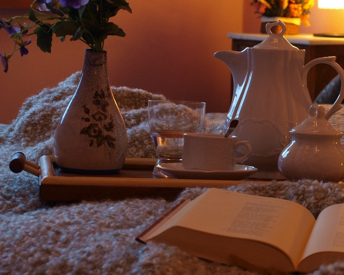 Textiles cálidos y naturales Tazón de café y libro sobre una manta, con flores y una tetera en un ambiente acogedor.